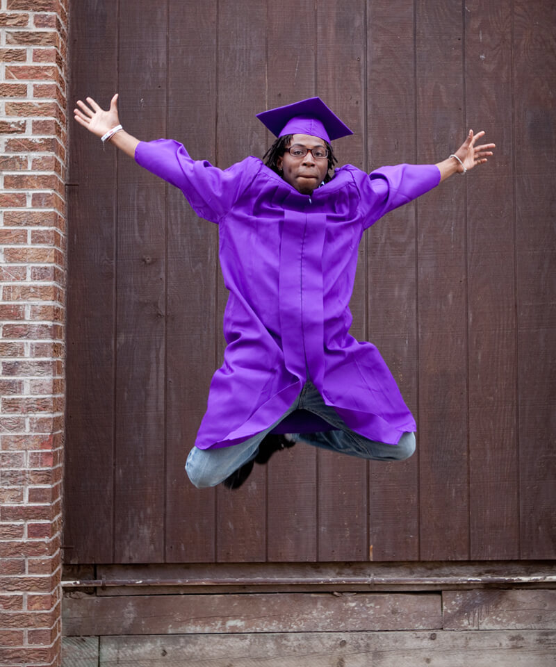 graduating student jumping in the air wearing a purple graduation cap and gown - Denver Street School